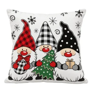 Cojín blanco con tres gnomos navideños de gorros rojos y negros, un árbol verde y un regalo, sobre fondo blanco.